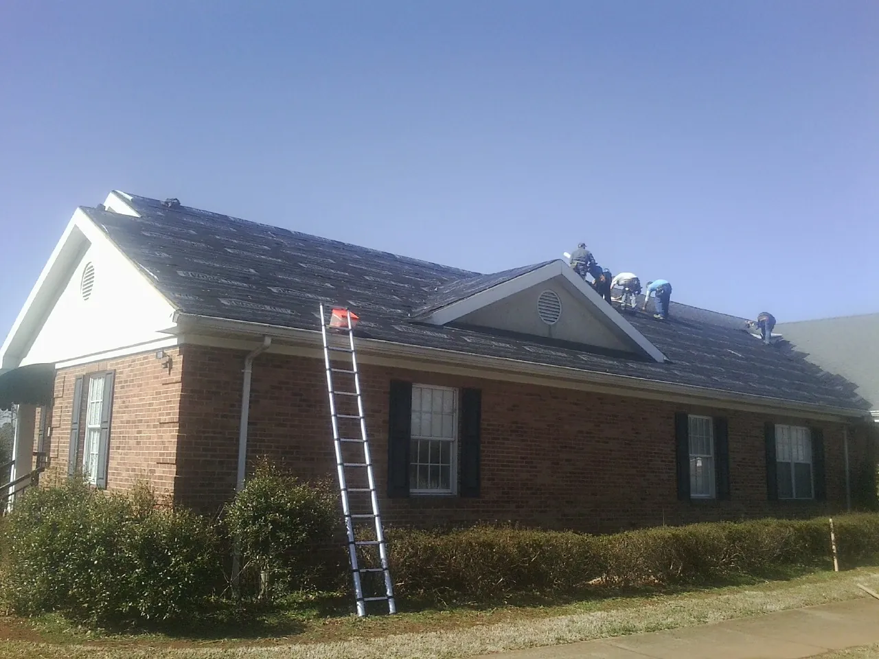 Crew tearing shingles off a brick ranch roof with a ladder set — Metal Roof Repair work by our crew in Volant