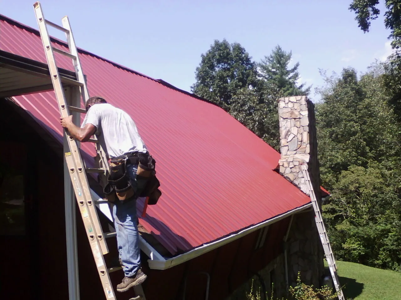 Roofer on a ladder beside a red metal roof and stone chimney — Soffit & Fascia Repair by Roofing Brothers in Volant